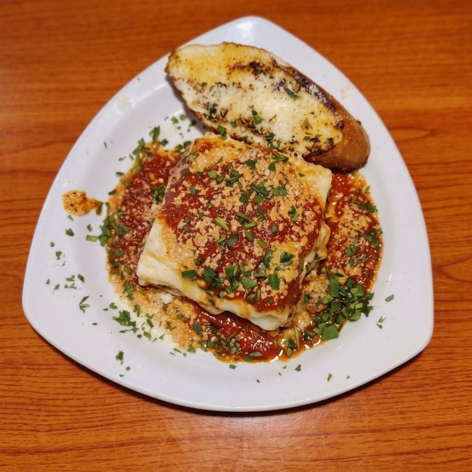 Layered lasagna with tomato sauce, breadcrumbs, and parsley served with garlic bread on a white plate.