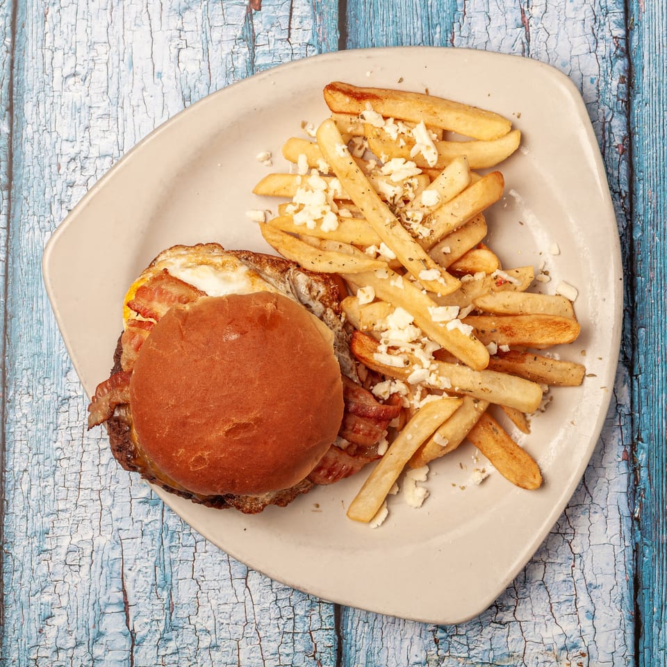Bacon cheeseburger with a fried egg alongside feta-topped seasoned fries on a plate over a blue weathered wooden table.