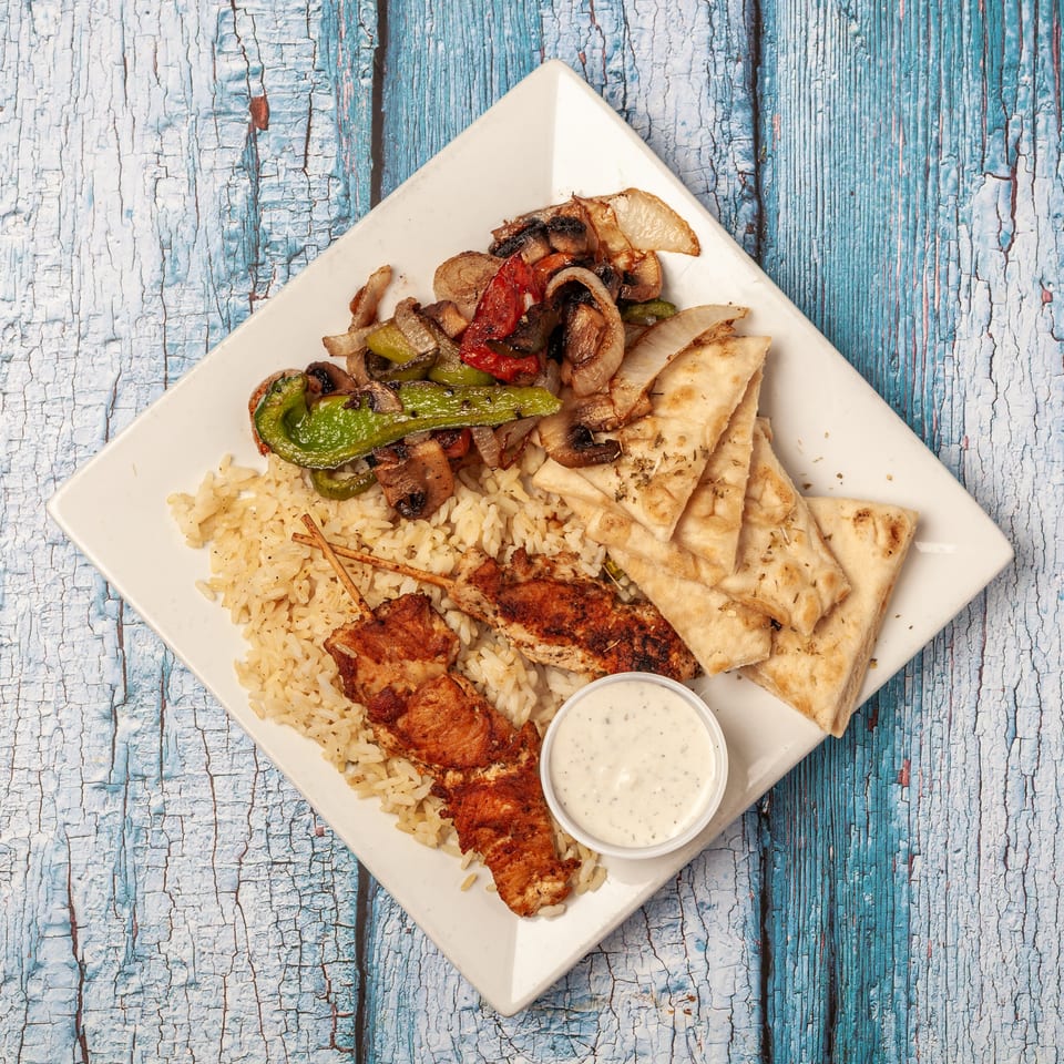 Mediterranean platter with grilled chicken, rice, sautéed vegetables, flatbread, and creamy dipping sauce on a white plate.