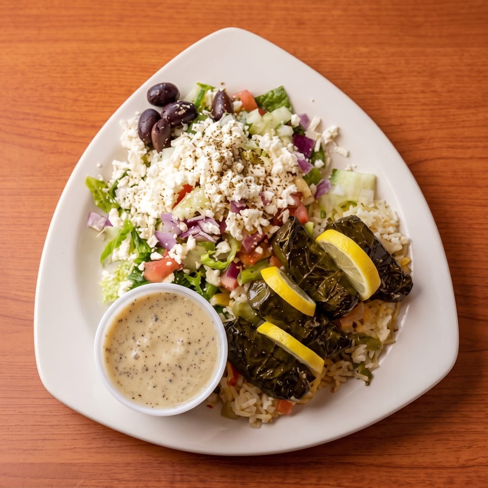 Greek salad with feta, olives, vegetables, rice, stuffed grape leaves, lemon wedges, and creamy dressing on a white plate.