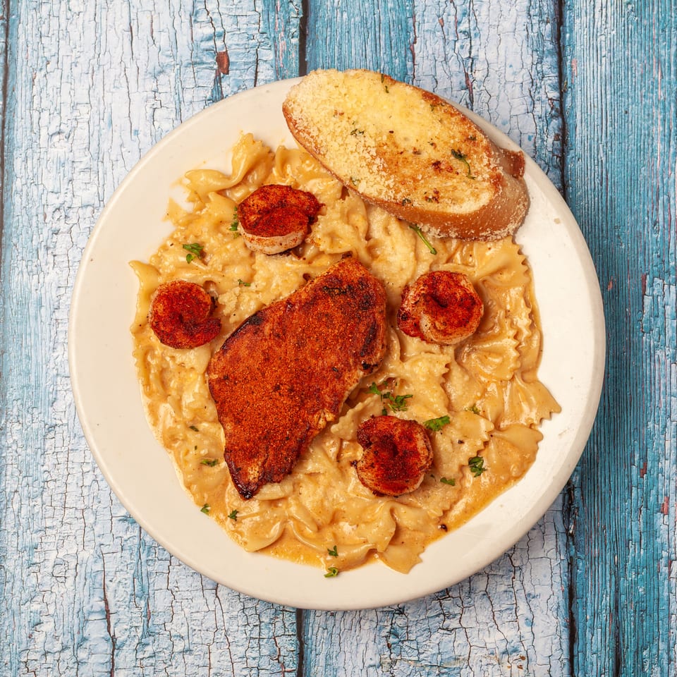 Creamy bow-tie pasta topped with seasoned grilled chicken, shrimp, parsley, and garlic bread on a white plate.